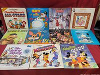 Overview of all 11 kids records albums displayed on a red surface showing different colorful illustrated covers featuring characters such as Charlie Brown, Frosty the Snowman, Disney characters, and Sesame Street's Ernie.