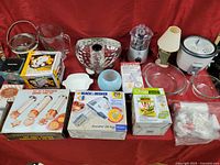 Overview of all items in the lot arranged on a red cloth backdrop including boxed kitchen appliances, lamps, glassware, and miscellaneous kitchen items.