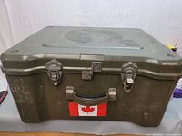 Front view of the green plastic military storage box showing metal latches, handle with Canadian flag sticker, and wear marks.