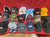 Overview of 17 diverse baseball caps showing various colors, logos, and styles laid out flat on a red surface.
