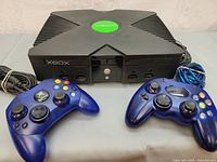 Front view of Microsoft Xbox console with two translucent blue wired controllers placed in front, showing general condition and details of console and controllers.