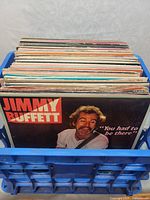 Blue plastic milk crate filled with stacked vinyl record albums showing Jimmy Buffett album cover in front.