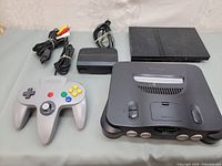 Photo showing Nintendo 64 console, Nintendo 64 grey controller, Nintendo 64 power supply adapter, AV cables, and Sony PlayStation 2 console in the background.