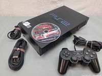 Sony PlayStation 2 slim console with game disc on top and bundled cables including power and AV cable beside the console, along with a wired Sony controller visible.