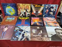 Wide shot showing 12 of the rock and other genre vinyl album covers arranged on red cloth surface.