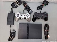Sony PlayStation 2 console, two wired DualShock 2 controllers (silver and black), power adapter, AV cable laid out together on white surface.