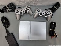 Top-down photo of silver Sony PS2 console, two silver DualShock 2 controllers, power and AV cables.