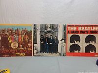 Three vinyl albums by The Beatles propped side by side showing front covers: Sgt. Pepper's Lonely Hearts Club Band, Hey Jude, and A Hard Day's Night.