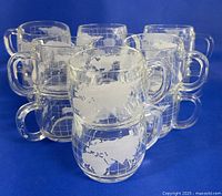 Group of twelve clear glass mugs arranged in a semi-circle, showing etched world map designs covering the entire mug body with detailed continental and grid pattern in white.