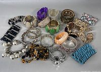 Photo showing over 20 different bracelets and cuff bangles arranged on a white surface. The collection includes various styles such as metal cuffs, beaded bracelets, faux pearl strands, and wide resin bangles in different colors.