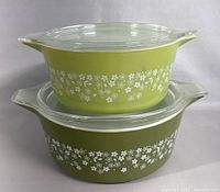 Two vintage PYREX nesting bowls stacked with lids, larger olive green bowl at the bottom and smaller light green bowl on top, both featuring white floral pattern.