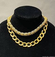 Two gold-tone necklaces displayed on a black velvet stand; one floral chain and one large link chain.