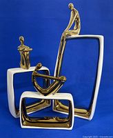 Front view of three modernist ceramic sculptures with abstract gold-tone figures on white rectangular bases against blue background