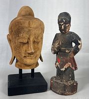 Front view of the carved Buddha head on stand with the painted temple figurine beside it, showing the facial details and pose.