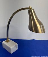 Desk lamp showing marble base and curved brass gooseneck arm with brass lampshade against blue background.