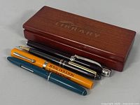 Wooden pen case labeled 'THE LIBRARY' with four pens placed on top: yellow Royal Doulton Lambethware pen, black pen, teal pen, and dark maroon pen.