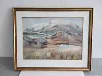 Framed watercolor painting showing a mountain landscape with water, grass, and rocks.
