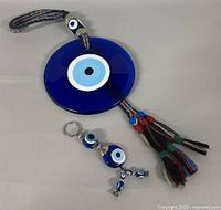 Photo of the blue glass evil eye wall hanging ornament with braided multicolored cord and tassels, alongside the blue evil eye keychain with beads and charms.