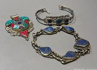 Photo showing old silver pendant, bracelet with blue stones, and turquoise black accent bangle cuff all placed on a grey surface.