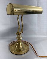 Front right angle view of vintage brass piano bankers desk lamp showing polished brass curved shade, adjustable dual arm, and weighted round base with orange cord.
