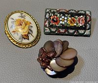 Three vintage French brooches displayed together on a gray surface including a Limoges porcelain oval brooch, a rectangular micro mosaic floral brooch, and a layered mother-of-pearl brooch.