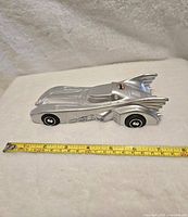Side view of a silver diecast Batmobile model placed on a white surface with a measuring tape showing about 7.5 inches in length.