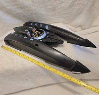 Side angle view of the black Batboat toy with seated Robin figure, placed next to a measuring tape showing length.