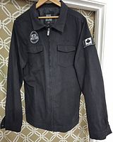Front view of black Metal Mulisha men's jacket showing two chest pockets with flaps, zipper closure, logo patch on chest, and Canadian flag patch on sleeve.