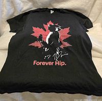Full front view of black T-shirt with Tragically Hip design featuring singer silhouette, red maple leaf, and 'Forever Hip.'