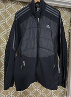 Front and full view of men's Adidas Terrex hybrid light jacket, black with synthetic fill quilted panels and breathable fabric parts, Adidas stripes on shoulders and sleeves, Terrex logo on front pocket.