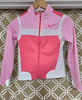 Front view of pink Nike girls athletic jacket on hanger showing color block design with white accents and front zipper.