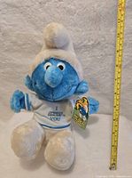 Front view of a blue Smurf plush doll wearing a white hat, white pants, and a white shirt embroidered with 'I SMURF YOU.' The doll has the original Smurf character tag attached to its arm.