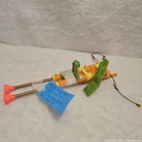 Photo of double barrel plunger toy gun laid on white textured fabric, showing side view with yellow and green parts and two orange plungers extended.