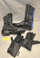 Side and upright view of black leather mid-calf women's motorcycle boots with side zippers, paired with black fingerless gloves with decorative white embroidery.