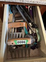 Top view inside kitchen drawer showing assorted kitchen utensils including wooden meat claws, spatulas, tongs, pizza cutter, and other tools.