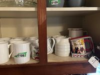 Shelf with assorted coffee and tea mugs including plain white mugs, Lyngby mug with berry illustration, ribbed ceramic mug, and holiday-themed mug with red handle.