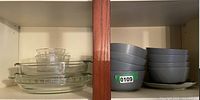 Photo showing 3 clear glass pie plates stacked on left shelf and 8 gray ceramic cereal bowls stacked on the right shelf with one small saucer plate underneath.