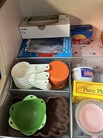 Photo showing measuring cups, silicone lids, funnels, brown silicone mold, paper cupcake liners, and Reynolds baking cups organized in wire trays.