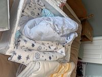 Photo of bedding sets and blankets piled in a basket, showing a white and blue snowflake patterned blanket and white striped bedsheet