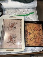 Two breakfast trays on a mattress showing different decorative designs, one floral and bird themed tray and one vintage world map tray