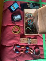 Photo showing multiple bracelets including beaded, braided fabric, and metal bangles in a wooden box and laid out on a red surface.