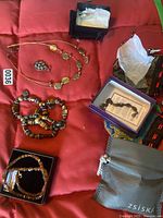 Photo showing several costume jewelry items on a red fabric background, including a layered necklace with gold-tone disks, a beaded bracelet, earrings, a pendant in a purple box, and a branded drawstring pouch.