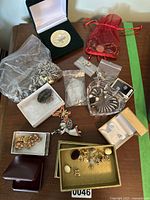 Overview of costume jewelry on a wooden surface including multiple items in plastic bags, small boxes, and a red pouch. Includes silver chain necklace, brooches, and a round silver coin in black display case.