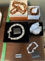 Four pieces of costume jewelry on a wooden table including Joan Rivers Classics Collection pearl necklace and bracelet in original boxes, and two amber-tone beaded strands; one coiled and one laid out.