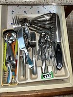 Top view of a kitchen drawer containing assorted stainless steel forks, spoons, knives, plastic-handled kitchen utensils including a ladle, a peeler, and measuring spoons. A large chef's knife with black handle is placed on the tray.