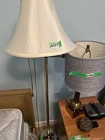 Floor lamp with tall brass base and white fabric shade with a flared shape, photographed indoors near a nightstand and other items.