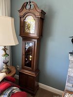 Full view of Ridgeway solid wood grandmother clock standing next to a lamp and furniture, showing its tall and elegant form.