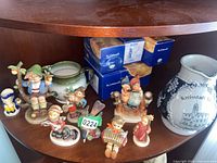 Shelf shot showing eight assorted M.J. Hummel figurines and a large decorative ceramic jug with German text and building illustration, plus original boxes.