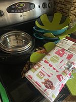 Photo showing stacked cookware including a large black pan, turquoise roasting/fry pans, green drying mats, a stainless steel mesh drying rack and kitchen towels.