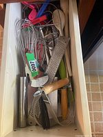 Drawer with assorted kitchen utensils including graters, whisks, peelers, wooden spoons, and other tools.